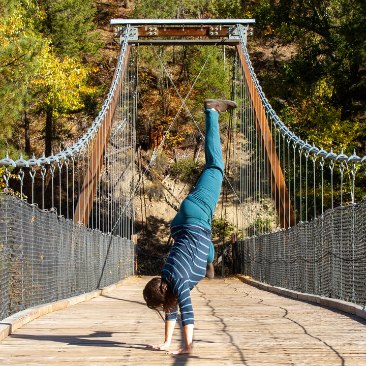 A person doing a handstand on a wooden suspension bridge with trees in the background.