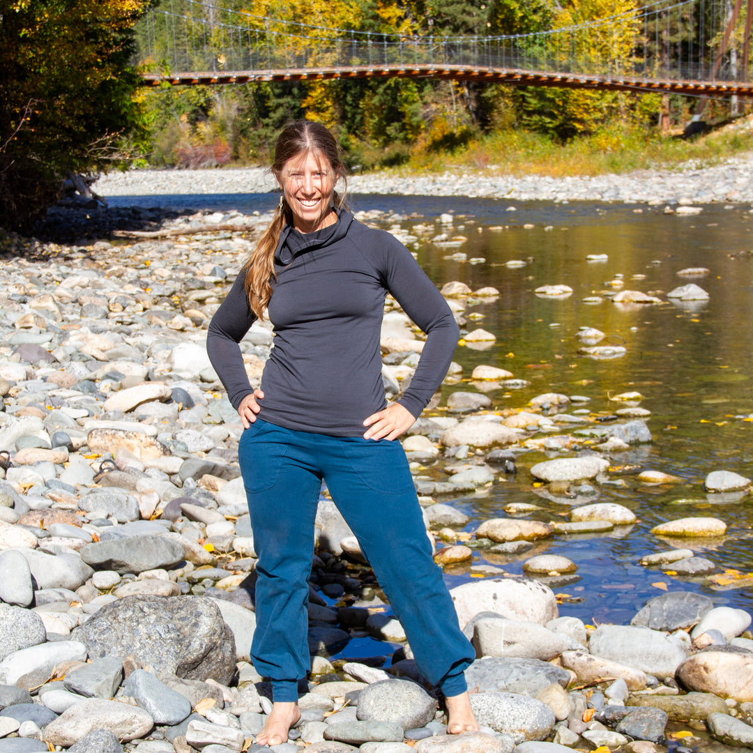 A woman stands barefoot on a rocky riverbank outdoors, wearing a dark charcoal long-sleeve top and teal pants, with water, trees, and a suspension bridge in the background.