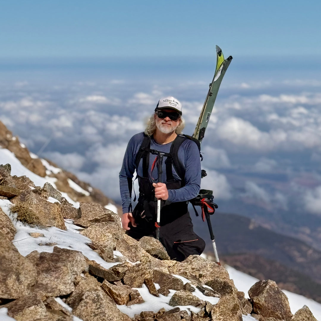 Man with skis on a mountain peak with a scenic view of clouds below wearing a merino wool base layer shirt